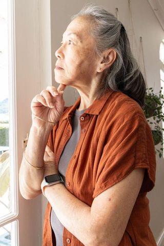 Senior asian woman at window contemplating wearing rust shirt and smartwatch in daylight