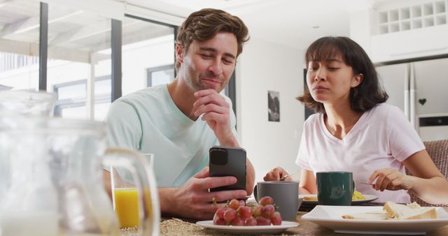 Casual morning scene with young couple smiling while checking a smartphone at breakfast table. Various foods and drinks visible, including grapes, toast, and coffee. Ideal for content regarding relationships, technology use in daily life, morning routines, and healthy living.