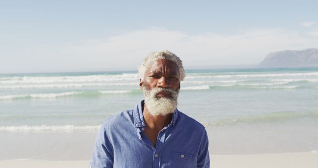 An elderly man with gray hair and a white beard stands on a sandy beach with waves gently crashing in the background. The man is wearing a casual blue shirt, and the scene conveys a sense of peace and tranquility. This image is ideal for content related to leisure, retirement, travel, senior lifestyle, wellness, and nature.