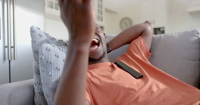 Man relaxing on couch in home while having a joyful conversation on smartphone. Spacious living room in background. Suitable for themes related to leisure, home life, modern communication, and happiness.