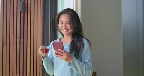 Smiling asian woman holding smartphone and glass of tea by window, relaxed home lifestyle