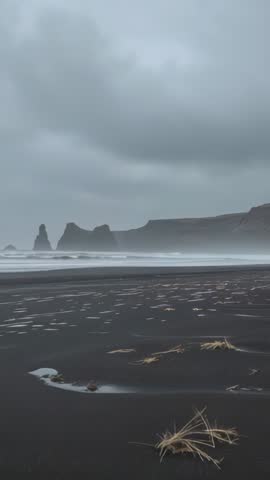Vertical video of waves crashing on black sand beach with windblown grass and sea stacks