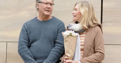 Mature couple sharing warm conversation while holding bouquet by beige stone facade