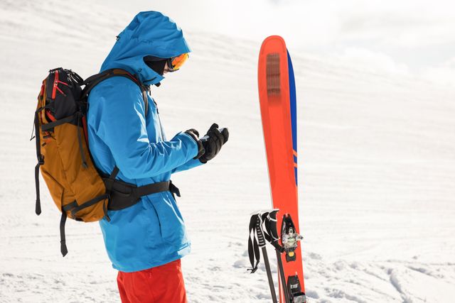 Skier standing on snowy mountain using mobile phone, wearing blue jacket and orange backpack. Ideal for content related to winter sports, outdoor activities, adventure travel, technology in nature, and active lifestyles.
