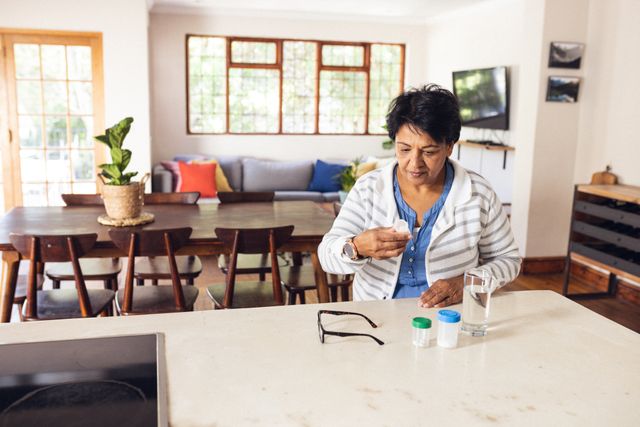 Biracial mature woman with eyeglasses, water and drug bottles on table taking medicines at home. Copy space, short hair, unaltered, healthcare, pills, sickness, treatment and retirement concept.