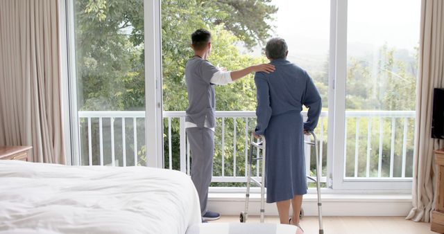 Shows caregiver assisting senior woman using walker by large window. The caregiver's hand is on the senior's shoulder, providing support and comfort. Use for themes of caregiving, elderly support, wellness, healthcare services, home care assistance, patient rehabilitation, and aiding mobility or balance in the elderly. Ideal for healthcare and senior care advertisements, brochures, websites, and articles highlighting elderly well-being.