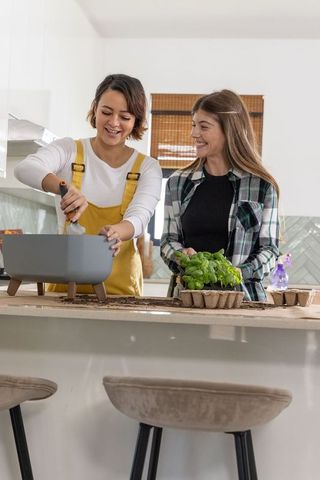 Two women potting basil at kitchen island wearing mustard overalls and plaid shirt, smiling
