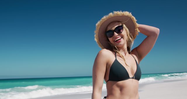 Young woman wearing sunglasses and a sunhat enjoying a sunny day at the beach. She is smiling and appears relaxed and carefree, dressed in a black bikini. The bright blue sea and sky provide a vibrant backdrop. Ideal for use in travel promotions, summer fashion advertisements, lifestyle blogs, and social media campaigns focusing on vacation or beach activities.