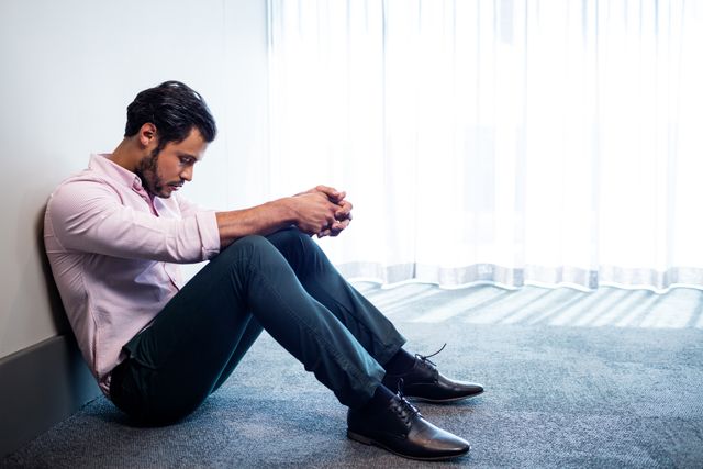 Businessman sitting against a wall in an office, looking down with a sad expression. This image can be used to illustrate themes of workplace stress, mental health issues, corporate burnout, and emotional challenges faced by professionals. Suitable for articles, blogs, and campaigns focused on mental health awareness and support in the workplace.