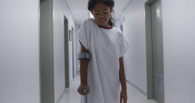 Young girl wearing medical gown using crutch in hospital hallway shows healthcare scenario and recovery process. Suitable for topics on pediatric care, medical facilities, injury recovery, and healthcare services.
