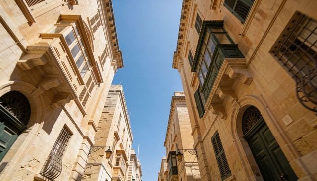 Sunlit narrow limestone alley with green oriel balconies pointing to blue sky