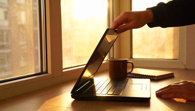 Hand lifting laptop lid on sunlit windowsill during golden hour warm workspace