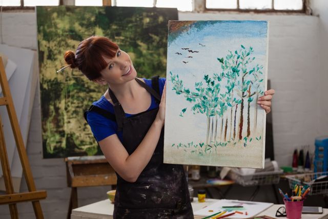 Young woman smiling and holding her painting of trees in an art class. Ideal for use in articles or advertisements related to art education, creative hobbies, art studios, and personal growth through artistic expression.
