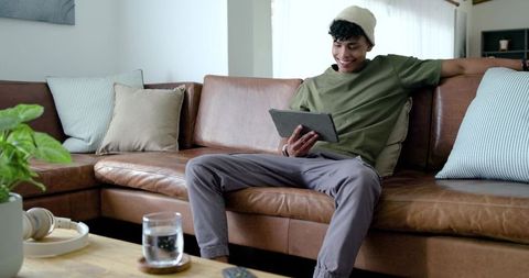 Young man relaxing on leather sofa holding tablet for streaming and remote work