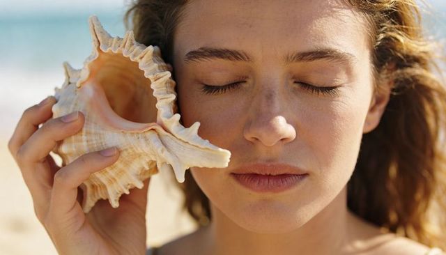 Adult woman listening to conch shell on sunny beach, serene freckled close-up
