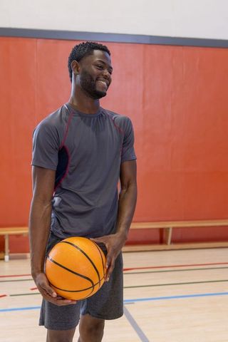 Smiling african american basketball player standing on indoor court holding ball near waist