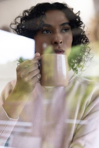 Contemplative non-binary adult drinking from pale pink mug by window with soft reflections