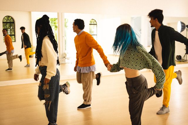 Group of diverse young dancers energetically practicing dance steps in modern dance studio. They are holding hands and maintaining balance while smiling and enjoying their session. Ideal for content related to fitness, cultural diversity, teamwork, lifestyle, and dance training tutorials.