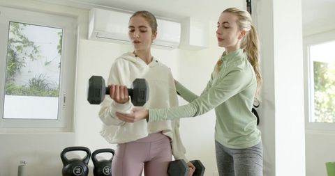 Female trainer guiding client lifting dumbbell in sunlit home gym for strength training