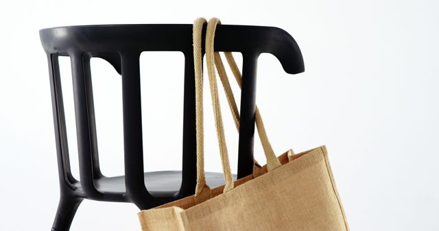The image showcases an environmentally friendly jute bag hanging on a stylish modern black chair. This is ideal for promoting sustainability, eco-friendly lifestyle choices, reusable products, or minimalist and modern interior designs. It can be utilized in blogs, websites, or marketing materials focused on environmental consciousness, sustainability, and stylish home decor.