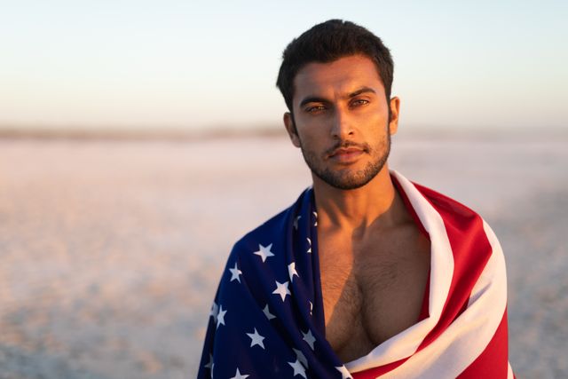 Young man wrapped in American flag standing on beach at sunset. Ideal for themes of patriotism, national pride, summer activities, and outdoor lifestyle. Suitable for use in advertisements, social media posts, and editorial content related to American culture and holidays.