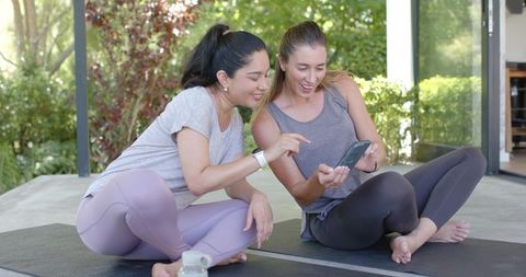 Two women on yoga mats sharing workout app, pointing at smartphone and laughing outdoors