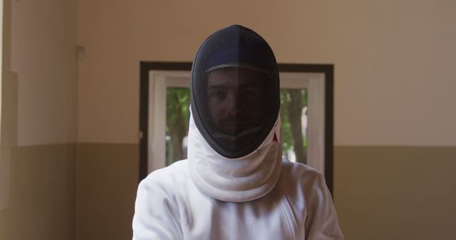 This visual shows a fencer wearing full fencing gear, including a protective mask and uniform, standing indoors before a competition or practice session. Suitable for use in content related to fencing, sports training, athlete preparation, combat sports, and protective gear. Ideal for illustrating dedication and focus in sports, training routines, and the importance of proper equipment in contact sports.