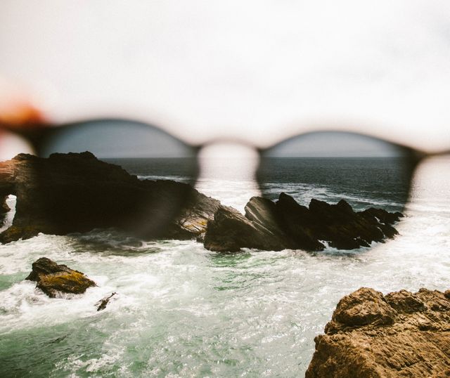 Clear, coastal scene highlighting waves crashing against rock formations. Ocean visible through sunglasses, bringing attention to changed perspective from blurred foreground to sharp middle and background. Ideal for travel blogs, beach-themed promotions, advertisements for sunglasses or visual representation of perspective shifts and focus in presentations.