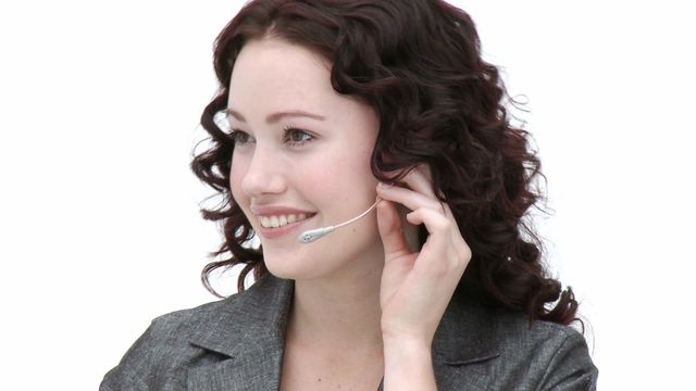 Smiling young woman wearing professional business attire with curly hair, working in a call center with a headset. Ideal for illustrating business service environments, customer service roles, or representing communication in promotional materials aimed at depicting professionalism in various industries.