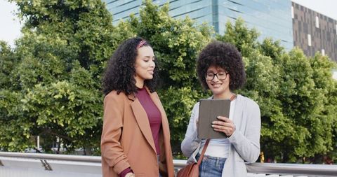 Diverse women smiling while sharing tablet and collaborating on urban bridge with greenery