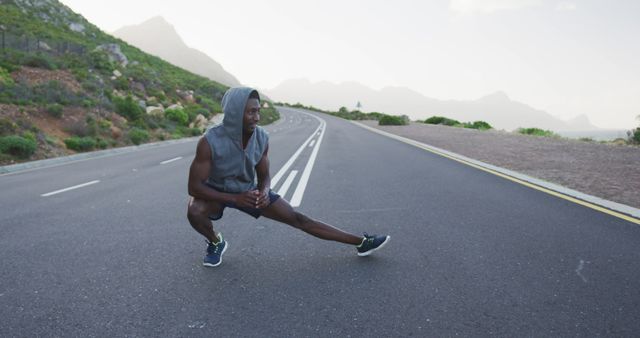 African american man wearing a hoodie performing stretching exercise on the road - Download Free Stock Photos Pikwizard.com