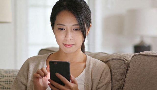 Woman using smartphone on cozy sofa tapping screen and relaxing in bright living room