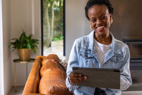 African american woman smiling while holding tablet near leather sofa in living room