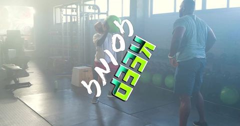 Man squatting holding green medicine ball while coach observing in backlit gym