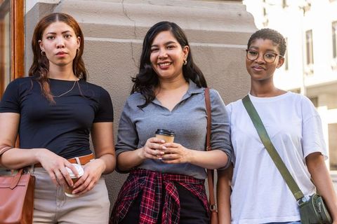 Three women standing on city street smiling and holding coffee cups