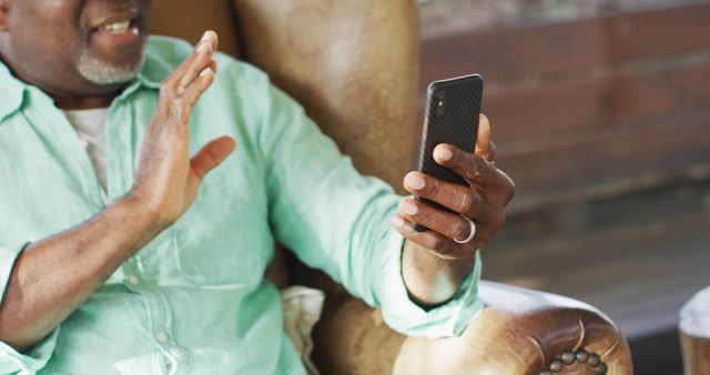 Happy african american senior man sitting in armchair making image call with smartphone, waving. retirement lifestyle, spending time alone at home.