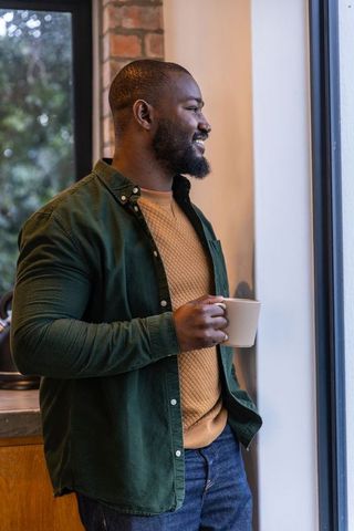 Cozy bearded african american man standing by window holding coffee mug smiling in kitchen