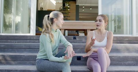 Two friends sitting and talking on modern house steps in activewear, sunlit porch chat