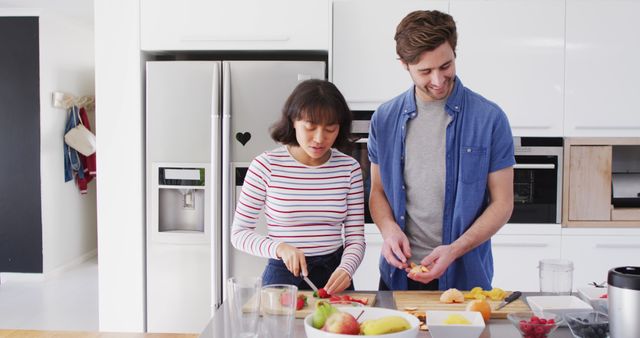 A couple who looks happy and engaged preparing a fresh fruit salad at home, symbolizing teamwork, domestic life, and healthy eating practices. This image can be used for advertisements and articles on healthy living, nutrition, cooking classes, or content focused on relationships and family life. It is ideal for websites, blogs, and marketing materials promoting kitchen appliances or recipe blogs.