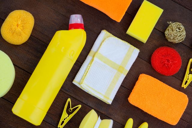 Cleaning supplies including a yellow bottle, cloth, various sponges and scrubbers are spread out on a wooden floor. This arrangement is perfect for use in advertisements for cleaning products, housekeeping services, and hygiene-related content, emphasizing cleanliness and order.