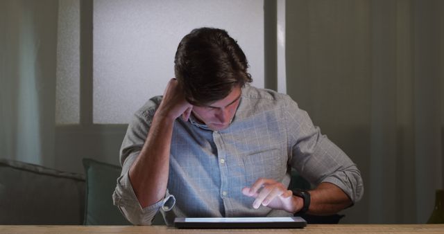 Young man intently using a tablet computer at home in a dark room. This image can be used to represent technology use, modern living, concentration, and home office setups. It is also suitable for illustrating concepts like working or studying at night, focus. Ideal for online articles, blogs, advertisements related to tech products, or social media content.