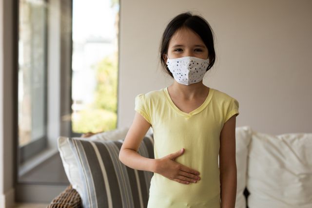 Little caucasian girl standing in front of the couch in their house while wearing a face mask. she has her hand on her stomach