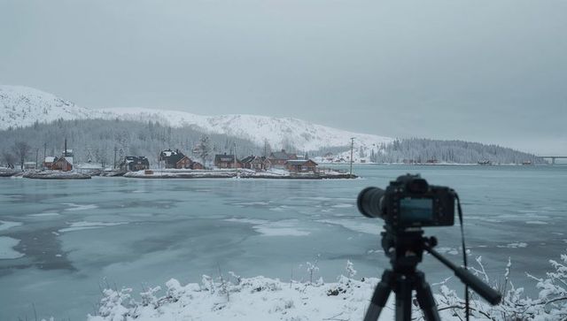 Camera on tripod capturing snowy nordic village across frozen inlet with telephoto lens