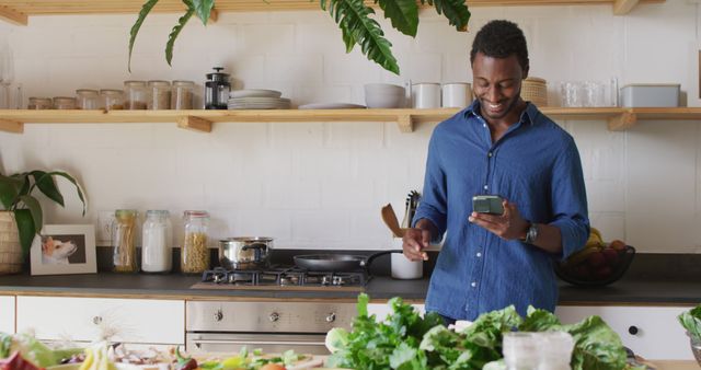 Man standing in modern kitchen, smiling and checking phone while cooking with fresh vegetables on counter. Perfect for themes of home cooking, lifestyle, and technology in daily life. Can be used for articles about modern kitchen designs, cooking apps, or balanced lifestyle.