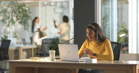 Indian woman working on laptop in modern coworking office with colleagues in background