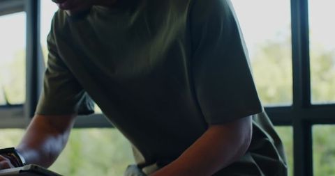 Man leaning over tablet at window interacting with device wearing olive tee and wristwatch