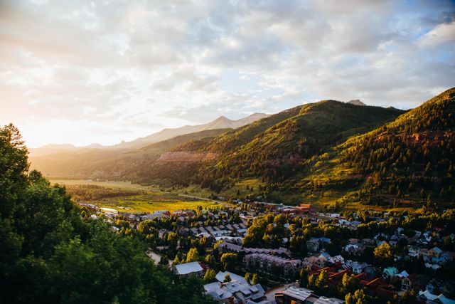 Morning sun illuminating mountain valley with a quaint village nestled below. Clear sky with scattered clouds enhances the picturesque beauty. Suitable for travel brochures, landscape photography collections, and nature-themed blogs.