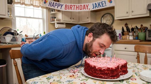Man leaning pressing face into pink birthday cake at kitchen table with banner and candles