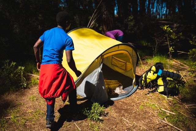 This image shows an African American couple setting up a yellow tent in a forest. They are surrounded by trees and greenery, indicating a natural, outdoor setting. The scene suggests teamwork and adventure, making it ideal for use in content related to camping, trekking, outdoor activities, travel, and nature exploration. It can be used in blogs, travel guides, advertisements for outdoor gear, and social media posts promoting adventure and outdoor experiences.
