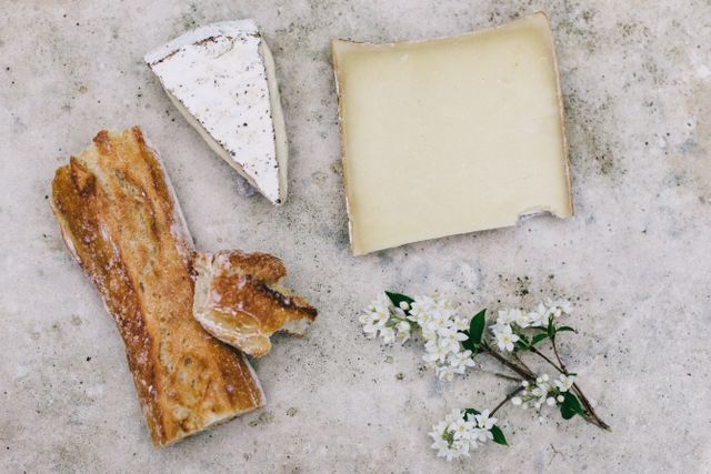 Assorted cheeses and pieces of baguette are arranged on a rustic surface alongside small white flowers. Ideal for use in culinary blogs, gourmet food websites, food styling inspiration, or advertisements for cheese retailers and bakeries.